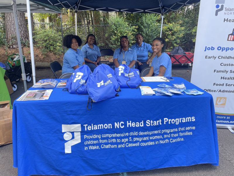 A group of five women wearing matching blue shirts sit behind a table covered with a blue tablecloth that reads “Telamon NC Head Start Programs.” The table displays brochures, blue drawstring bags with the Telamon logo, and informational materials. A banner to the right lists job opportunities, and the setting appears to be an outdoor community event under a canopy.