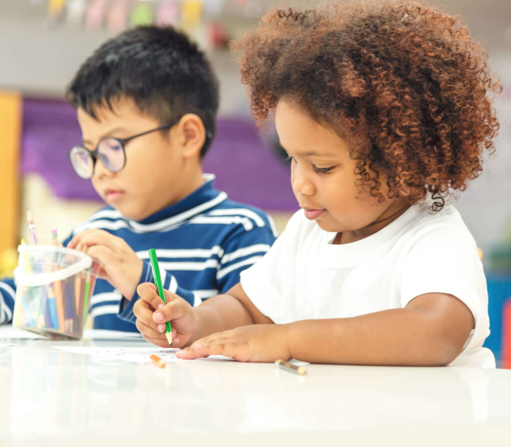 Two young children sitting at a table in a classroom, focused on coloring and drawing with colored pencils, with one child wearing a striped shirt and glasses and the other in a white t-shirt