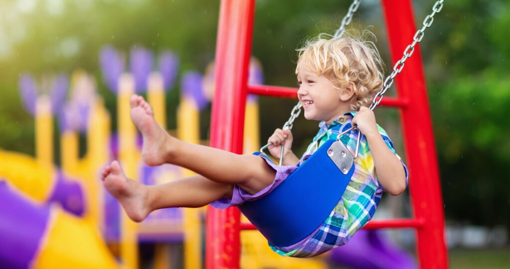 Smiling young child with blonde curly hair enjoying a swing ride at a colorful playground, wearing a plaid shirt and barefoot