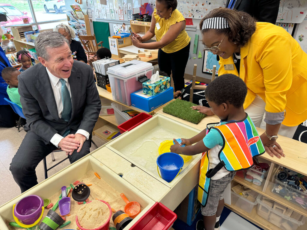 A man in a suit and a woman in a yellow jacket watch a young boy play at a sensory water and sand table in a classroom. The boy wears a colorful striped apron while scooping water with a toy. Other children and adults are visible in the background engaging in activities.