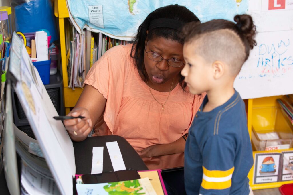 A teacher wearing glasses and a peach-colored top points at a large chart while speaking to a young boy standing beside her. They are in a colorful classroom with books, maps, and learning materials visible in the background. The teacher appears to be explaining something, and the boy listens attentively.