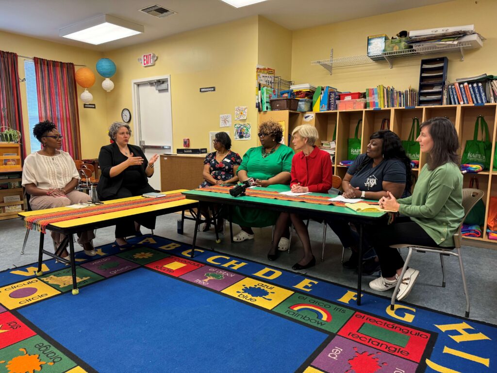 A group of six adults sit at tables in a classroom, engaged in a discussion. Colorful rugs, bookshelves, and hanging paper lanterns decorate the room.