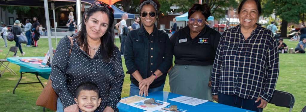A group of four women stand behind a table covered with a blue tablecloth that reads “Wake County Smart Start.” The table displays flyers and a children’s book. In front of the table, a smiling young boy holds a blue snow cone while standing with a woman who has her arm around him. The event takes place outdoors under tents with other people visible in the background.