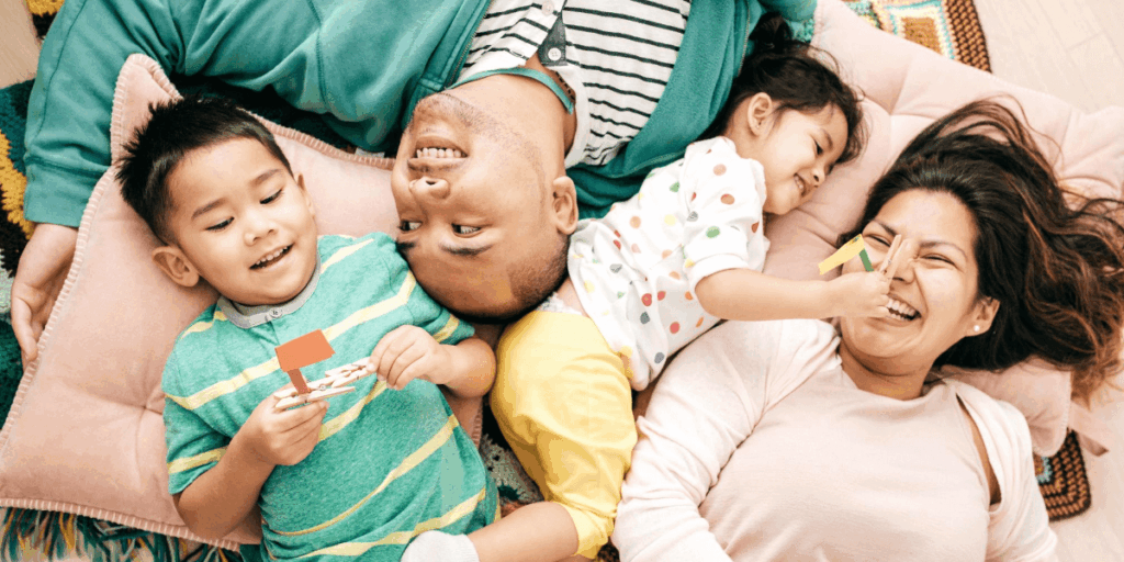 Happy family of four lying on the floor together, smiling and playing, with the young daughter playfully holding a clothespin on her mother’s nose