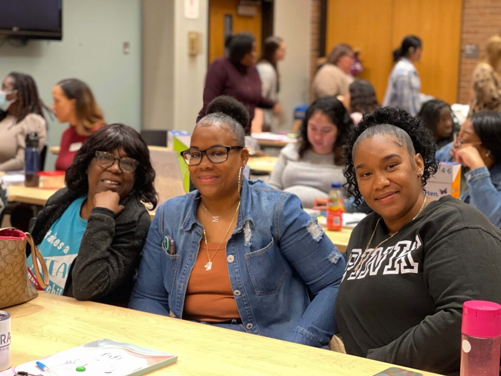 Three women sit together at a table during a professional development session, smiling at the camera.